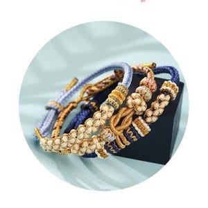 May include: Close-up of several braided bracelets in shades of blue, gold, and cream. The bracelets feature intricate knot and floral designs. The background is a soft, light blue, and the bracelets are displayed on a black surface.