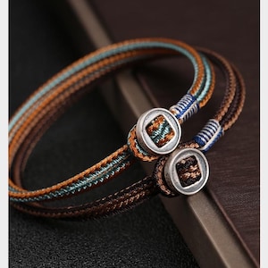 May include: Two braided bracelets with brown, orange, and teal threads. Each bracelet has a silver-colored clasp with a square design. The bracelets are displayed on a dark surface, showcasing the intricate details of the woven design.
