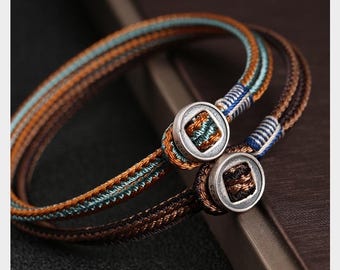 May include: Two braided bracelets with brown, orange, and teal threads. Each bracelet has a silver-colored clasp with a square design. The bracelets are displayed on a dark surface, showcasing the intricate details of the woven design.