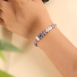 May include: A bracelet featuring a central lavender knot, flanked by white woven floral-shaped beads and small lavender spacers. The design is strung on a thin lavender cord, creating a delicate and intricate accessory with a soft color palette.