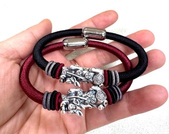 May include: Two bracelets featuring thick cord bands in black and deep red. Each bracelet includes a polished silver-toned metal cylinder clasp and a detailed silver-toned charm depicting a mythical creature adorned with circular coin-like patterns.