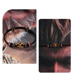 May include: A black cord bracelet featuring a central gold-toned knot ornament, accented with small gold beads and bands of red, white, and black thread. The adjustable design includes sliding knots to secure the fit around the wrist.