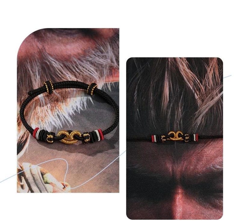 May include: A black cord bracelet featuring a central gold-toned knot ornament, accented with small gold beads and bands of red, white, and black thread. The adjustable design includes sliding knots to secure the fit around the wrist.