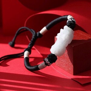 Op de afbeelding: Een zwart-wit gevlochten armband met een wit gesneden stenen middelpunt. De armband staat tegen een rode achtergrond, met een kleine rode kubus op de achtergrond. De armband is verstelbaar.