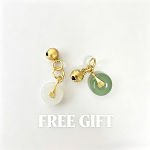 May include: Two gold-toned metal charms, each featuring a small spherical bell and a circular stone pendant. One pendant is translucent white, the other is sage green. Both are attached to gold jump rings. The text FREE GIFT is below.