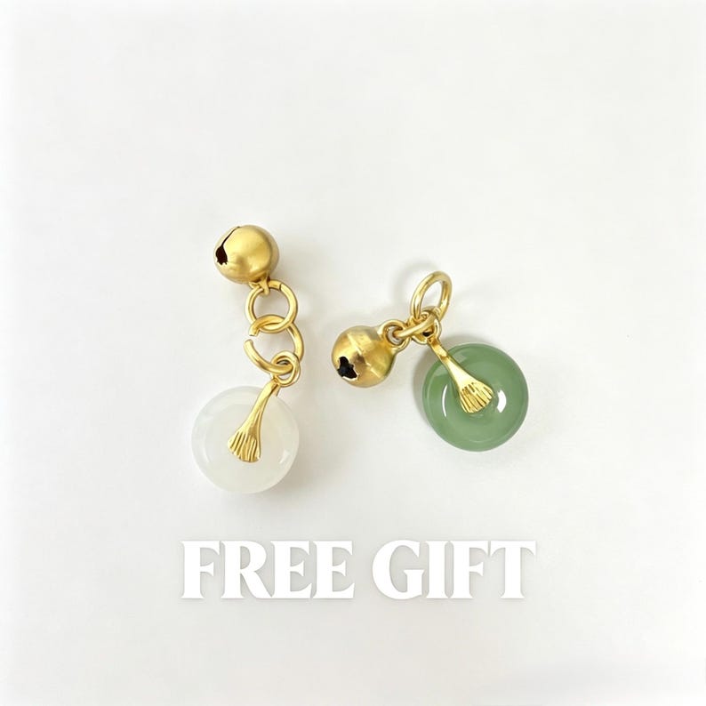 May include: Two gold-toned metal charms, each featuring a small spherical bell and a circular stone pendant. One pendant is translucent white, the other is sage green. Both are attached to gold jump rings. The text FREE GIFT is below.