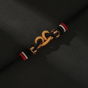 May include: A black bracelet with a gold-colored central design, featuring a chain-like pattern. The bracelet has red and white accents near the clasp. The background is a dark, textured surface.