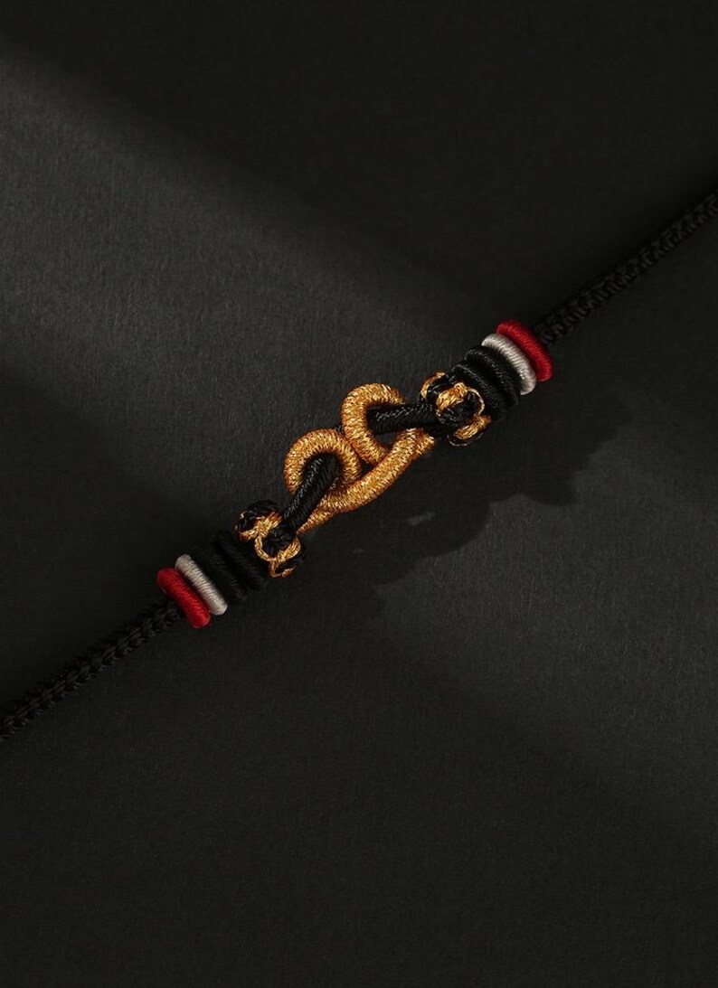 May include: A black bracelet with a gold-colored central design, featuring a chain-like pattern. The bracelet has red and white accents near the clasp. The background is a dark, textured surface.