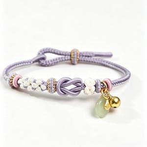 May include: A lavender cord bracelet featuring a central decorative knot, white braided accents, and small gold-toned beads. The bracelet includes a dangling charm set consisting of a smooth pale green stone and a small polished gold-toned bell.