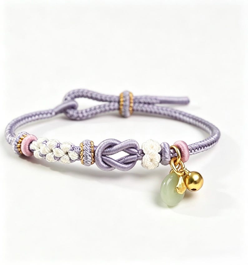May include: A lavender cord bracelet featuring a central decorative knot, white braided accents, and small gold-toned beads. The bracelet includes a dangling charm set consisting of a smooth pale green stone and a small polished gold-toned bell.