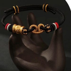 May include: A black braided bracelet with gold accents and a gold clasp. The bracelet features gold beads and a gold cylindrical charm. The bracelet has red and white accents. The bracelet is displayed on a dark surface.