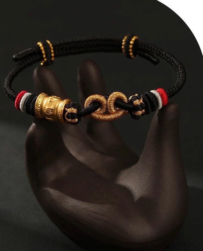 May include: A black braided bracelet with gold accents and a gold clasp. The bracelet features gold beads and a gold cylindrical charm. The bracelet has red and white accents. The bracelet is displayed on a dark surface.