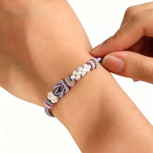 May include: A bracelet featuring a lavender cord base with a central knot design, accented by white braided segments, small pink circular beads, and metallic gold-toned spacers. The cord is tied in a secure knot, creating a layered, textured appearance.