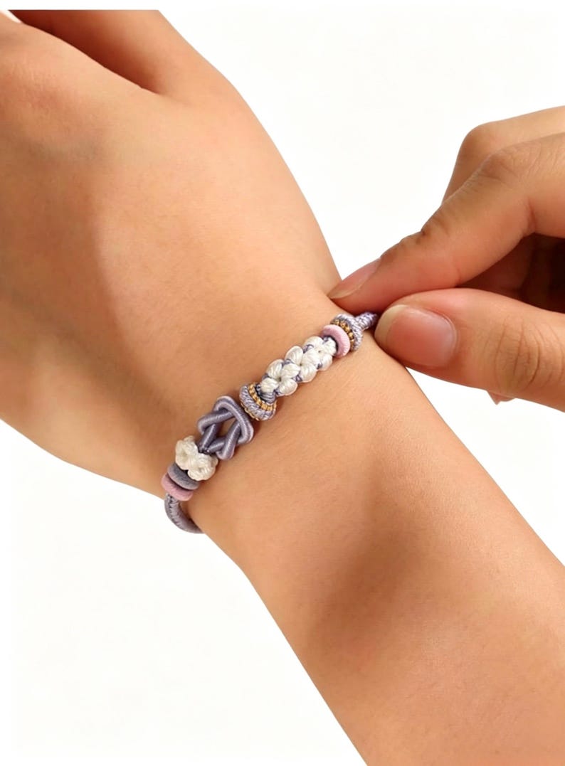 May include: A bracelet featuring a lavender cord base with a central knot design, accented by white braided segments, small pink circular beads, and metallic gold-toned spacers. The cord is tied in a secure knot, creating a layered, textured appearance.