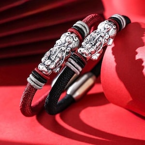 May include: Two bracelets with silver dragon head charms. One bracelet is red, black, and silver, the other is black, red, and silver. The bracelets are on a red surface.