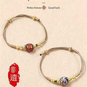 May include: Two bracelets with leather cords and lacquer beads. The beads are red, blue, and gold. The image text reads "Leather cord and lacquer bead bracelet", "Perfect fortune", and "Good luck". The length of the hand rope can be customised.