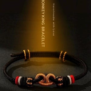 May include: A black cord bracelet featuring a central metallic gold knot, flanked by red and white thread accents. The bracelet includes small gold-toned beads and is inscribed with the text MONKEY KING BRACELET and Handmade with Love.