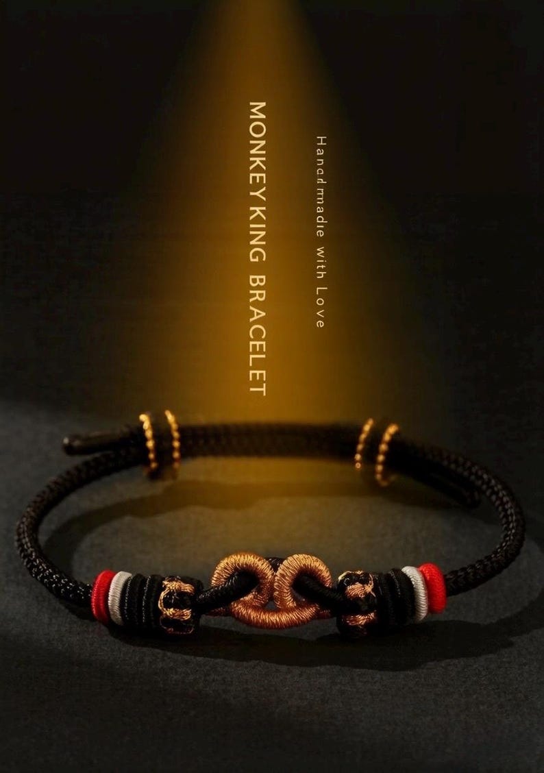 May include: A black cord bracelet featuring a central metallic gold knot, flanked by red and white thread accents. The bracelet includes small gold-toned beads and is inscribed with the text MONKEY KING BRACELET and Handmade with Love.