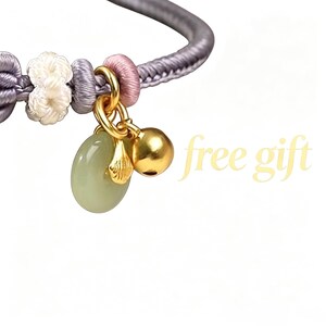May include: A bracelet featuring a grey braided cord with white and pink knotted accents. It includes a gold-toned bell charm, a small gold-toned shell-shaped charm, and a smooth, pale green oval stone charm. The text free gift is visible.