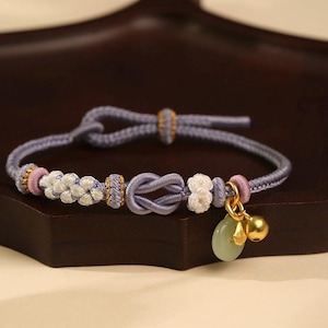 Puede incluir: Una pulsera de color lavanda con intrincados detalles de nudos y trenzas. La pulsera presenta detalles blancos y rosas, cuentas doradas y un pequeño dije ovalado verde con una campana dorada. Una delicada pieza de joyería.