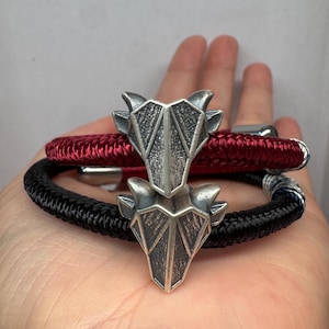 Puede incluir: Dos pulseras con dijes de cabeza de animal geométricos en tono plateado. Una pulsera está hecha de un cordón trenzado rojo oscuro y la otra de un cordón trenzado negro. Ambas pulseras incluyen terminales metálicos y un diseño angular texturizado.