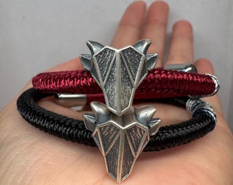 May include: Two bracelets featuring silver-toned geometric animal head charms. One bracelet is made of a deep red braided cord, while the other is made of a black braided cord. Both bracelets include metallic end caps and a textured, angular design.