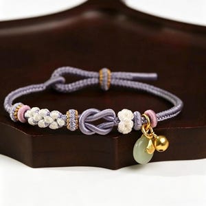 May include: A lavender cord bracelet featuring a central infinity knot, white woven floral accents, and small pink cylindrical beads. The bracelet includes a gold-toned bell charm and a smooth, pale green circular stone pendant on a gold ring.