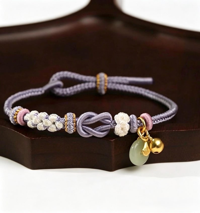 May include: A lavender cord bracelet featuring a central infinity knot, white woven floral accents, and small pink cylindrical beads. The bracelet includes a gold-toned bell charm and a smooth, pale green circular stone pendant on a gold ring.