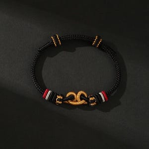 May include: Black bracelet with gold-colored accents. The bracelet features a braided black cord with gold-colored clasps and a gold-colored central design. Red and white accents are visible on the bracelet.