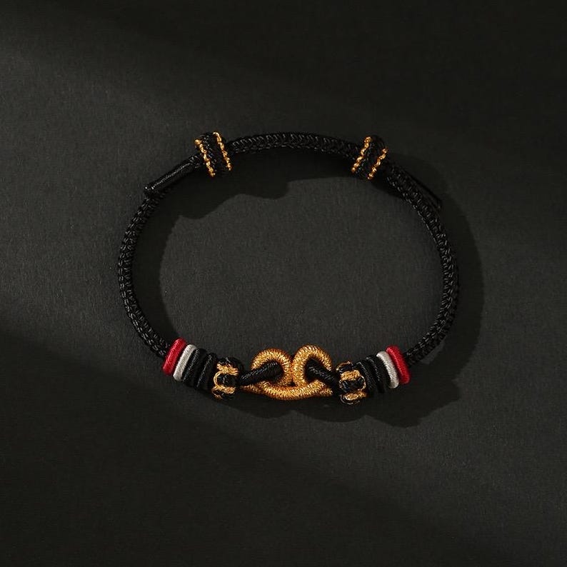 May include: Black bracelet with gold-colored accents. The bracelet features a braided black cord with gold-colored clasps and a gold-colored central design. Red and white accents are visible on the bracelet.