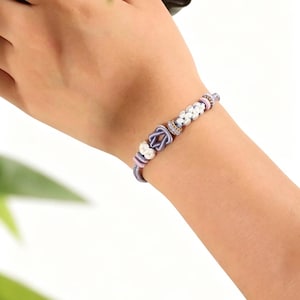 May include: A bracelet featuring a central lavender-colored cord tied into a knot, flanked by small white textured beads and smaller lavender and gold-toned spacers. The bracelet is constructed from a thin, braided cord in a soft lavender hue.