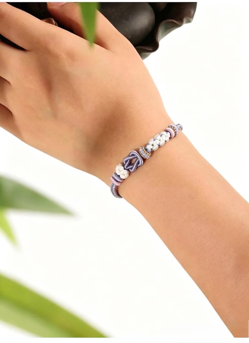 May include: A bracelet featuring a central lavender-colored cord tied into a knot, flanked by small white textured beads and smaller lavender and gold-toned spacers. The bracelet is constructed from a thin, braided cord in a soft lavender hue.