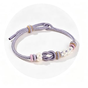 May include: A lavender cord bracelet featuring a central infinity knot, accented with smaller white and pink knotted segments and gold-toned beads. The bracelet includes an adjustable sliding knot closure made from the same lavender cord material.