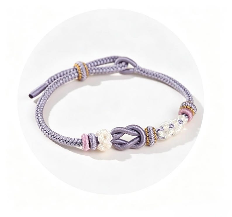 May include: A lavender cord bracelet featuring a central infinity knot, accented with smaller white and pink knotted segments and gold-toned beads. The bracelet includes an adjustable sliding knot closure made from the same lavender cord material.