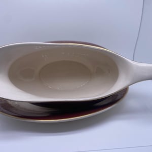 May include: A white gravy boat with a gold rim and a matching burgundy and gold patterned saucer. The gravy boat has a handle and a spout.