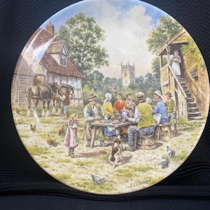 May include: A vintage ceramic plate with a colorful illustration of a group of people gathered around a table in a rural setting. The scene includes a church in the background, a horse-drawn cart, and a small cottage. The plate is signed "J.L.C. 1980".