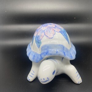 Könnte beinhalten: Eine kleine Porzellan-Schildkrötenfigur mit einem blauen und weißen Blumenmuster auf ihrem Panzer. Die Schildkröte ist in Blau- und Weißtönen bemalt, mit einer rosa Blume auf ihrem Rücken.