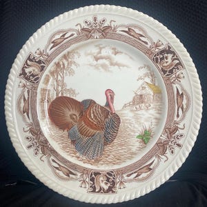 May include: A white ceramic dinner plate featuring a brown and white design of a turkey in a woodland setting. The plate has a rope-like border.