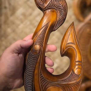 May include: A polished brown wooden fish hook carving featuring intricate spiral and linear relief patterns. The top section includes a circular iridescent shell inlay. The surface displays a smooth, glossy finish with deep wood grain textures.