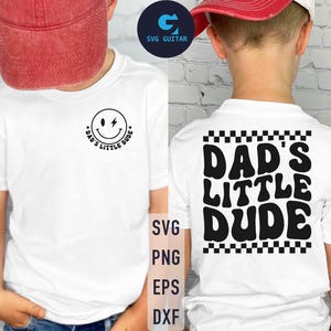 May include: Two white t-shirts with different designs. One shirt has a smiley face with a lightning bolt and the text "Dad's Little Dude". The other shirt has the text "Dad's Little Dude" in a retro font with a checkered pattern.