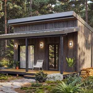 May include: A small, modern cabin with dark vertical wood siding and a slanted metal roof. The structure features a covered front porch with a white chair, glass sliding doors, a glass entry door, and three exterior wall-mounted light fixtures.