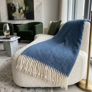 May include: A blue throw blanket with a woven herringbone pattern and long, cream-colored fringe along the bottom edge. The textile features a textured weave and is draped over a curved, off-white textured chair in a living space.