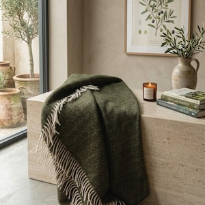 May include: A dark olive green throw blanket featuring a woven herringbone pattern and long, cream-coloured fringe along the edges. The textile is draped over a surface, showcasing its textured weave and the contrast between the green and cream.