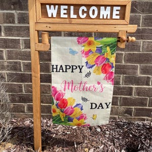 May include: A wooden welcome sign with a banner that reads "Happy Mother's Day." The banner features colorful flowers and butterflies. The sign is made of light brown wood and is placed outdoors.
