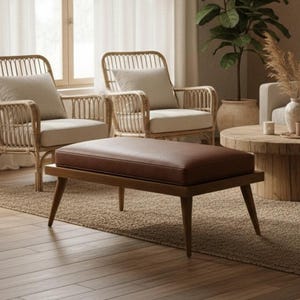 May include: A living room scene with neutral tones. Two rattan armchairs and a matching ottoman with a brown leather top are in the foreground. A light-colored sofa, round wooden coffee table, and decorative shelves complete the space. A large plant and sheer curtains add to the natural aesthetic.