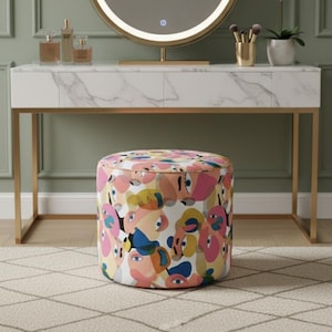 May include: A dressing table with a white marble top, gold legs, and a circular illuminated mirror. A patterned stool with pink, blue, and yellow shapes is in front. A white orchid and makeup brushes are on the table.