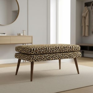 Puede incluir: Un otomán con estampado de leopardo y patas de madera oscura. El asiento rectangular de dos niveles está tapizado con un estampado de animales marrón y negro. El otomán está colocado sobre una alfombra beige claro en una habitación con una paleta de colores neutros.