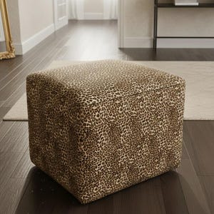 May include: Square ottoman with a leopard print pattern in shades of brown and tan. The cube-shaped furniture piece is placed on a dark wood floor. The ottoman is a decorative accent for a living room or bedroom.