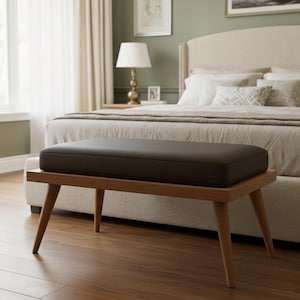 May include: A brown upholstered bench with a rectangular cushion sits on a wooden frame with angled legs. The bench is in a bedroom setting, with a bed and other furniture visible in the background.