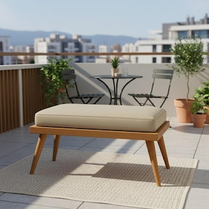 Pode incluir: Banqueta de madeira com almofada bege, colocada sobre um tapete de cor clara. A banqueta tem pernas anguladas e uma almofada retangular. O fundo mostra uma varanda com mesa, cadeiras e plantas em vasos.